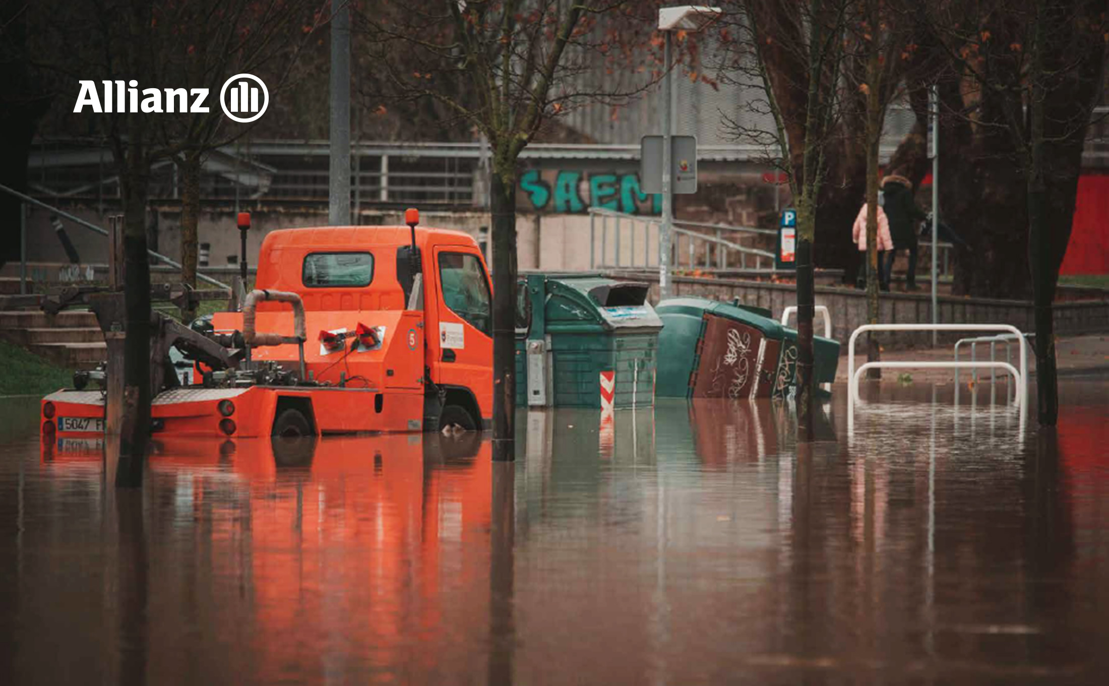 Flooded urban street with standing water covering the roadway and partially submerging an orange utility/tow truck. The white Allianz logo appears in the upper-left corner.