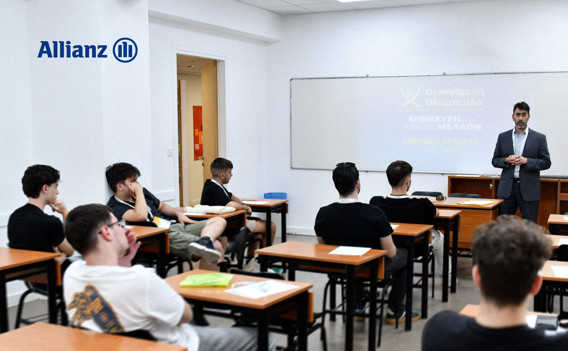 Άτομο κάνει παρουσίαση σε αίθουσα διδασκαλίας μπροστά σε ομάδα μαθητών που κάθονται σε θρανία. Στον τοίχο διακρίνεται λογότυπο της Allianz, ενώ πίσω από τον ομιλητή υπάρχει λευκός πίνακας και προβολή παρουσίασης σχετικής με την Ελληνική Οικονομική Ολυμπιάδα. Φωτεινός εσωτερικός χώρος εκπαιδευτικού χαρακτήρα.