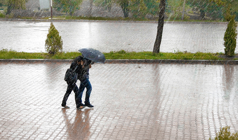 Two people walk across a rain-soaked paved area under an umbrella during heavy rainfall.